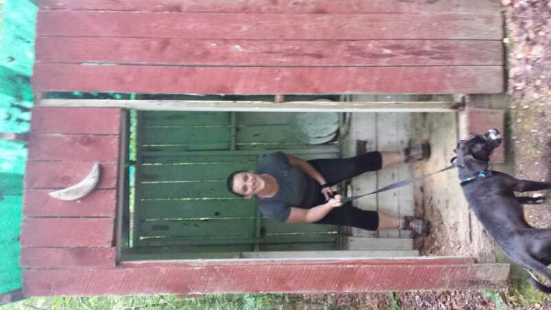 A person smiling while sitting inside a rustic wooden structure with a green interior, accompanied by a black dog on a leash. The exterior features red wooden planks and a decorative crescent shape. The setting appears to be outdoors, surrounded by greenery. Cedarville State Forest mountain bike trail.