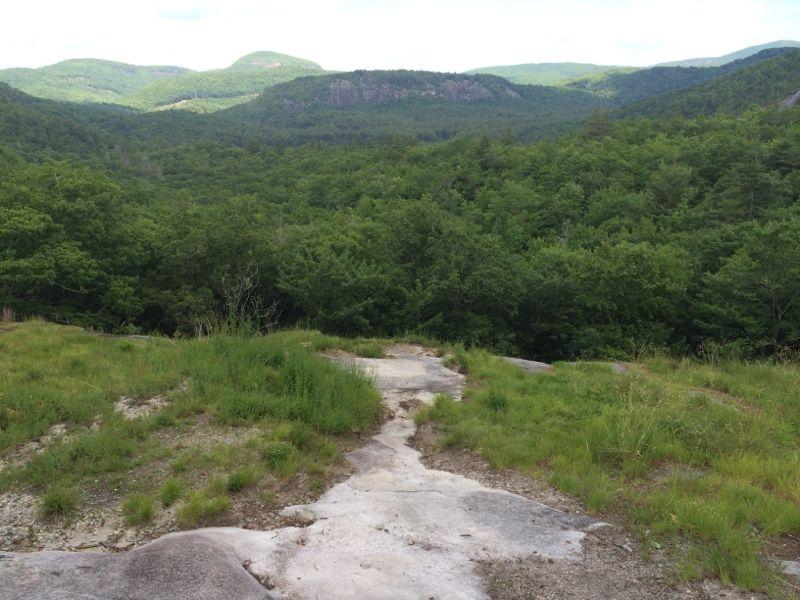 A scenic view of rolling green hills and dense forest under a partly cloudy sky, with a rocky pathway leading into the landscape. Panthertown Valley mountain bike trail.