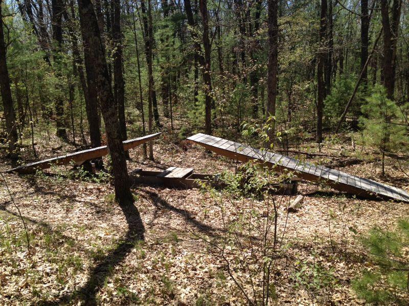 Wooden ramps situated in a forested area, surrounded by trees and scattered leaves on the ground. The ramps are unevenly placed, suggesting use for activities like biking or hiking. Sunlight filters through the branches, illuminating the natural setting. Hartman Creek State Park mountain bike trail.