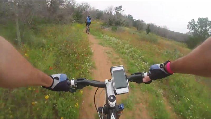 A person mountain biking on a dirt trail surrounded by wildflowers and greenery, with a smartphone mounted on the bike's handlebars. Another cyclist is visible in the background, riding along the path. Reveille Peak Ranch mountain bike trail.