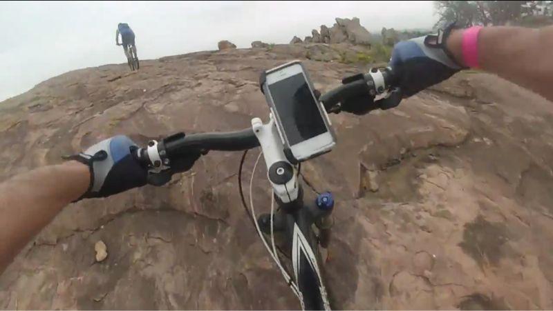 A cyclist's view from the handlebars of a mountain bike, showing rocky terrain ahead. The cyclist is wearing gloves and a watch, and a smartphone is mounted on the bike. In the background, another cyclist can be seen riding on the rocks. The scene is outdoors, suggesting a rugged biking environment. Reveille Peak Ranch mountain bike trail.
