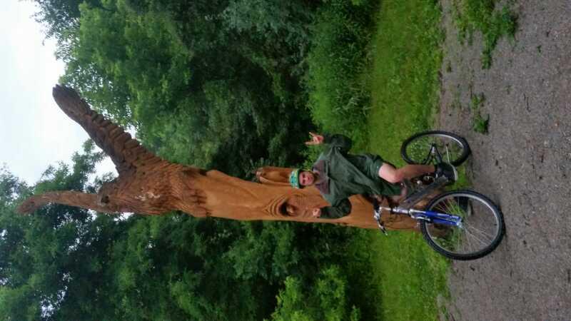 A person wearing a green rain jacket and a blue cap sits on a bicycle beside a large wooden sculpture of a hand raised in a gesture. The background features lush greenery, suggesting an outdoor park setting. Morton-Taylor Trail mountain bike trail.
