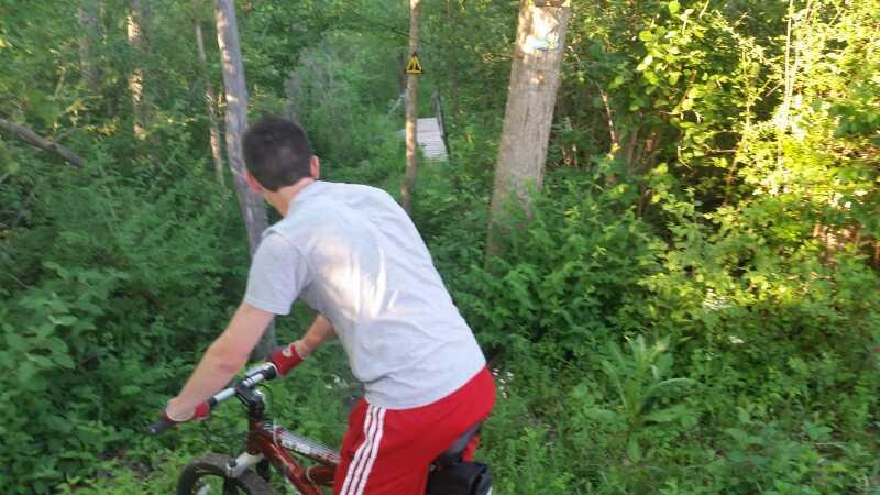 A person on a bike positioned on a wooded trail, looking down a slope towards a path. The scene is surrounded by greenery, with trees and bushes visible, and a caution sign in the background. The individual is wearing a gray shirt and red shorts. Morton-Taylor Trail mountain bike trail.