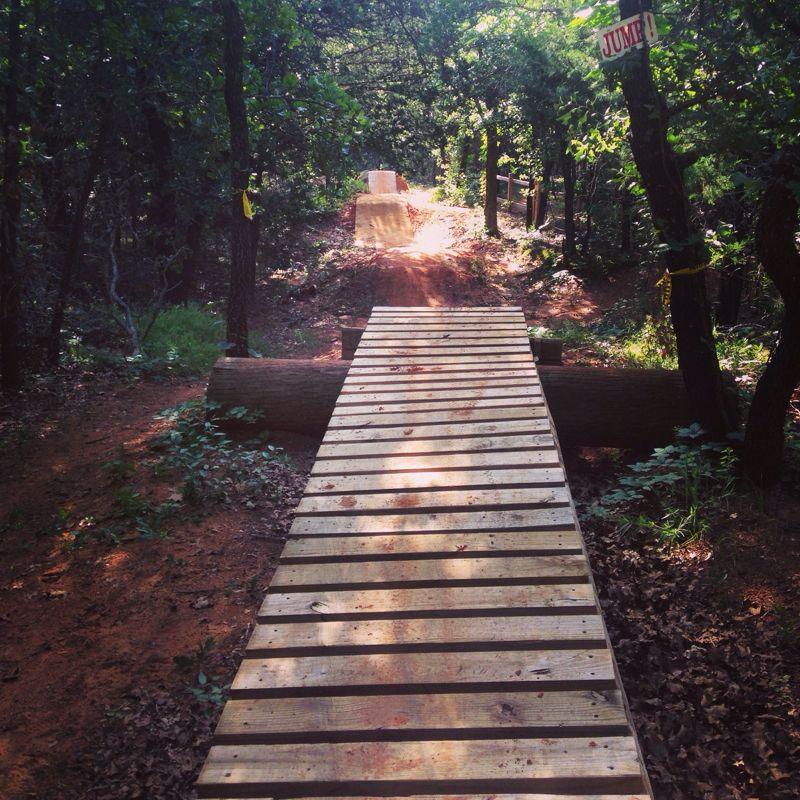 A wooden bridge leading to a dirt bike ramp in a wooded area, with trees surrounding the path. A sign on a nearby tree reads "JUMP!" indicating the ramp's purpose for biking or jumping. The ground is covered with brown dirt and scattered leaves, suggesting an outdoor recreational space. Lake Stanley Draper mountain bike trail.
