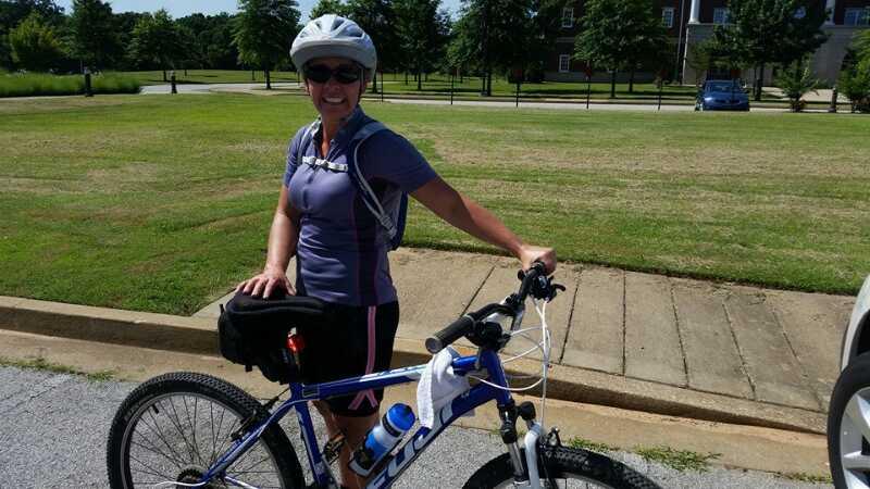 A woman in a helmet smiles while standing next to her blue mountain bike. She is wearing a purple athletic shirt and black shorts, with a backpack on her shoulders. The background features a grassy area and a parked car. Lake Ouachita Vista Trail (LOViT) mountain bike trail.