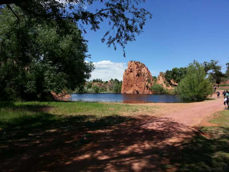 A scenic view of a tranquil lake surrounded by lush greenery and towering rock formations under a bright blue sky, with a sandy path leading to the water and people walking in the distance. Red Rock Canyon mountain bike trail.