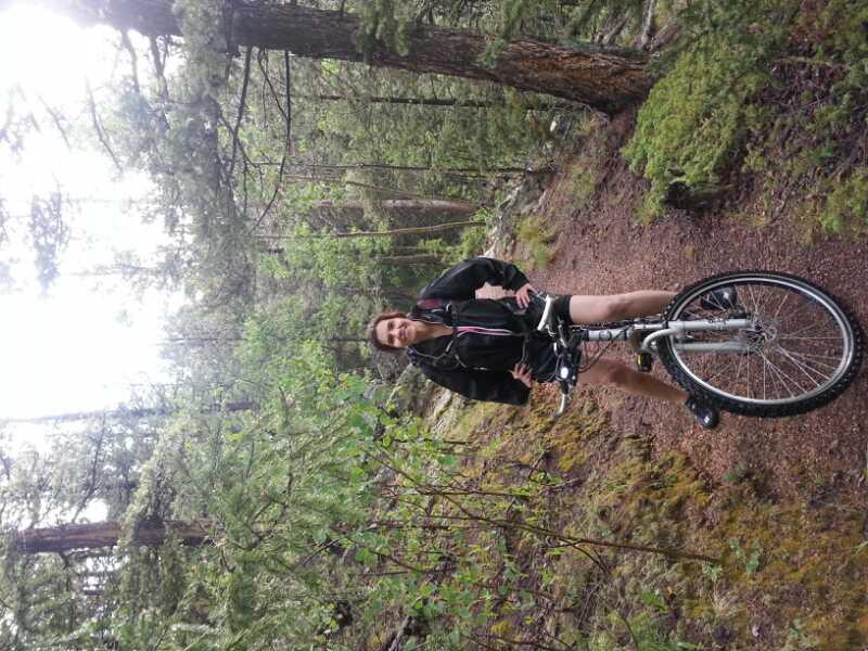 A person standing next to a mountain bike on a dirt path surrounded by lush green trees in a forest. The individual is wearing a black jacket and shorts, and is smiling, enjoying the outdoor environment. Rampart Reservoir mountain bike trail.