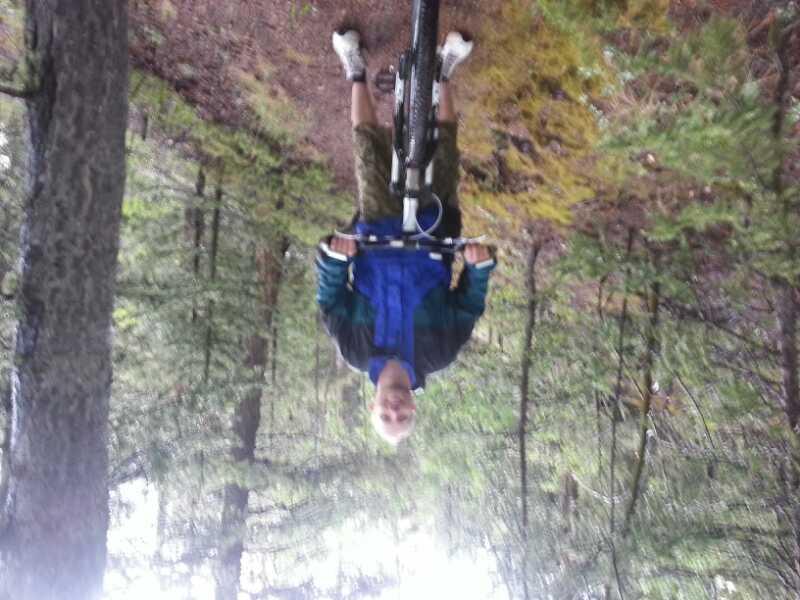 A person standing next to a mountain bike on a forest trail, surrounded by trees and greenery. The individual is wearing a blue jacket and shorts, and is facing the camera with a smile. Rampart Reservoir mountain bike trail.