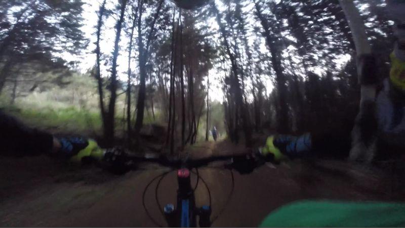 Alt text: A first-person view of mountain biking through a dense forest trail, with handlebars and gloves visible in the foreground. The path is narrow and surrounded by tall trees, while two people can be seen in the distance along the trail. The lighting suggests it is either early morning or late afternoon. DH Santa Elena - La Meseta - Medellin (Loreto) mountain bike trail.