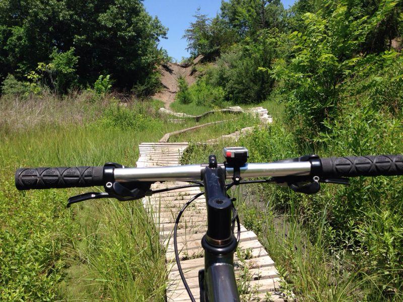 A mountain bike viewed from the handlebars, positioned on a narrow wooden path through lush, green vegetation. In the background, a steep hill rises, surrounded by trees and grass under a bright blue sky. The scene suggests a serene outdoor trail perfect for biking. Banner Lakes At Summerset Park mountain bike trail.