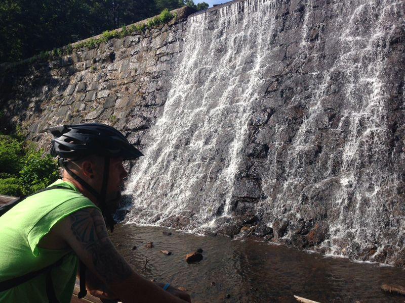 A person wearing a helmet sits beside a flowing waterfall, with a stone wall in the background. The scene captures the natural beauty of the waterfall surrounded by greenery and rocks. Paris Mountain State Park mountain bike trail.