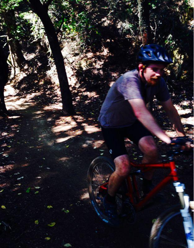 A person wearing a helmet and cycling attire rides a mountain bike along a dirt trail in a wooded area. Sunlight filters through the trees, casting dappled shadows on the ground. The cyclist appears focused and determined as they navigate the path surrounded by nature. El Corte De Madera Creek Open Space mountain bike trail.