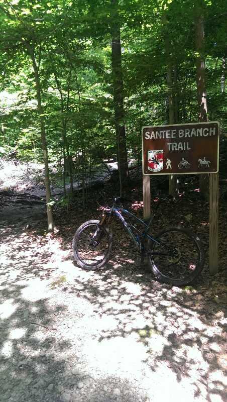 A mountain bike leaning against a wooden sign that reads "Santee Branch Trail," surrounded by lush green trees in a forested area. The trail seems to be a mix of dirt and gravel, with dappled sunlight filtering through the leaves. Patapsco Valley State Park (Avalon Area) mountain bike trail.