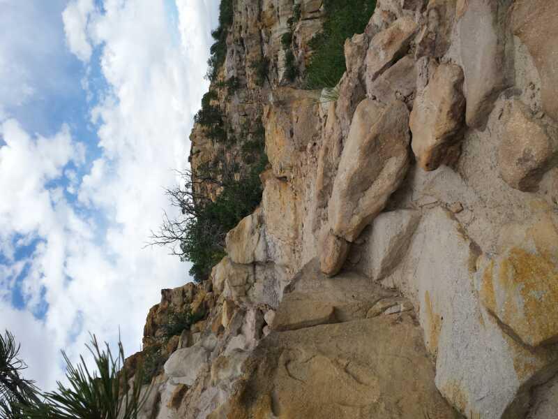 Rocky terrain with a steep cliffside, scattered boulders, and patches of greenery against a cloudy sky. Palmer Park mountain bike trail.