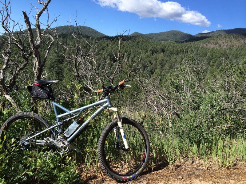 A mountain bike is parked beside a scenic view of rolling green hills and forested mountains under a blue sky with scattered clouds. The bike is positioned in the foreground with dry brush and grass surrounding it, creating a tranquil outdoor atmosphere. Cheyenne Mountain State Park mountain bike trail.