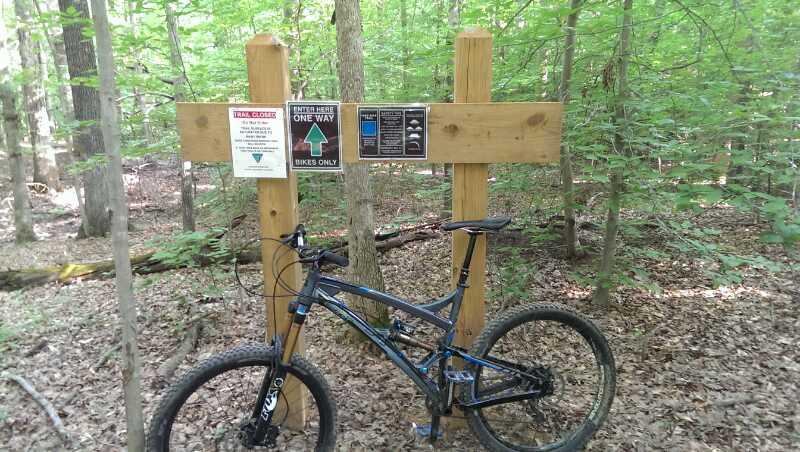 A mountain bike parked next to a wooden trail sign in a dense forest. The sign features multiple notices, including rules for trail usage, a directional arrow indicating a bike-only path, and guidelines for trail colors. The surrounding area is lush with greenery and fallen leaves. Meadowood mountain bike trail.