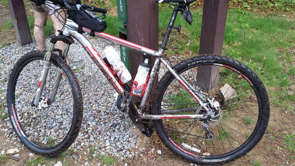 Trek Wahoo: A close-up image of a muddy mountain bike resting on gravel, with water bottles attached to the frame. The bike shows signs of recent use, with mud splattered on the frame and tires. In the background, a wooden signpost is visible, along with greenery.