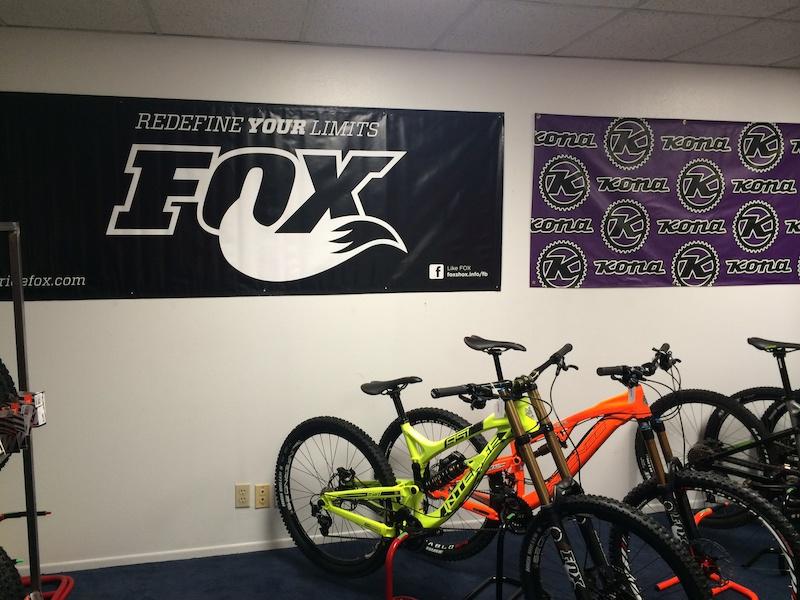 A display area featuring multiple mountain bikes in various colors, including a bright green bike and an orange bike. The wall is adorned with banners showcasing the FOX and Kona brands, with the FOX banner prominently stating "REDEFINE YOUR LIMITS." The overall setting appears to be a bike shop or showroom.