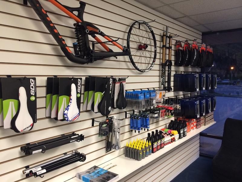 Alt text: A display wall in a bike shop featuring various bicycle components and accessories, including bike frames, saddles, tools, lubricants, and maintenance products arranged neatly on white slatwall.