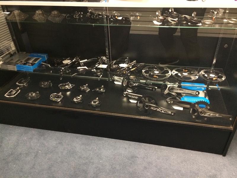 A display case showcasing various bicycle components, including pedals, cranksets, and other accessories. The items are arranged on a black shelf with a glass front, set against a neutral background.