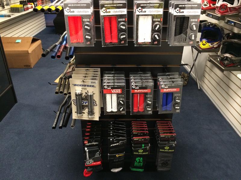 Display of various bicycle grips and accessories in a shop. The image shows a rack with packaged grips in red, white, black, and blue, labeled with brand names, alongside additional bike accessories and socks. The background features bike handlebars and helmets, indicating a cycling shop environment.