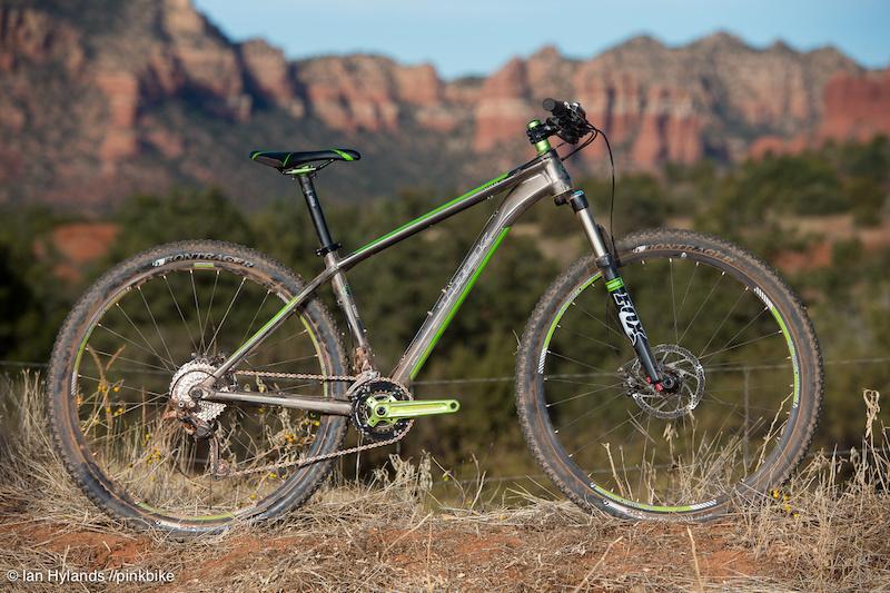 Trek Stache 8: Mountain bike positioned on a rocky terrain with a scenic backdrop of red rock formations and greenery. The bike features a sleek design with a silver frame and green accents, showcasing its wheels and gearing system.