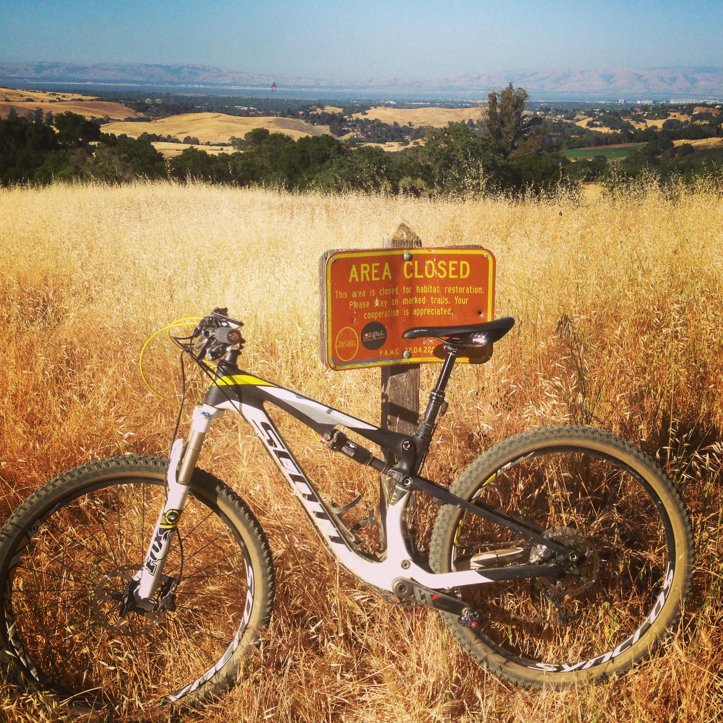 A mountain bike rests beside a closed area sign in a golden, grassy landscape with rolling hills in the background. The sign informs that the area is closed for habitat restoration and advises visitors to stay on marked trails. Arastradero Preserve mountain bike trail.