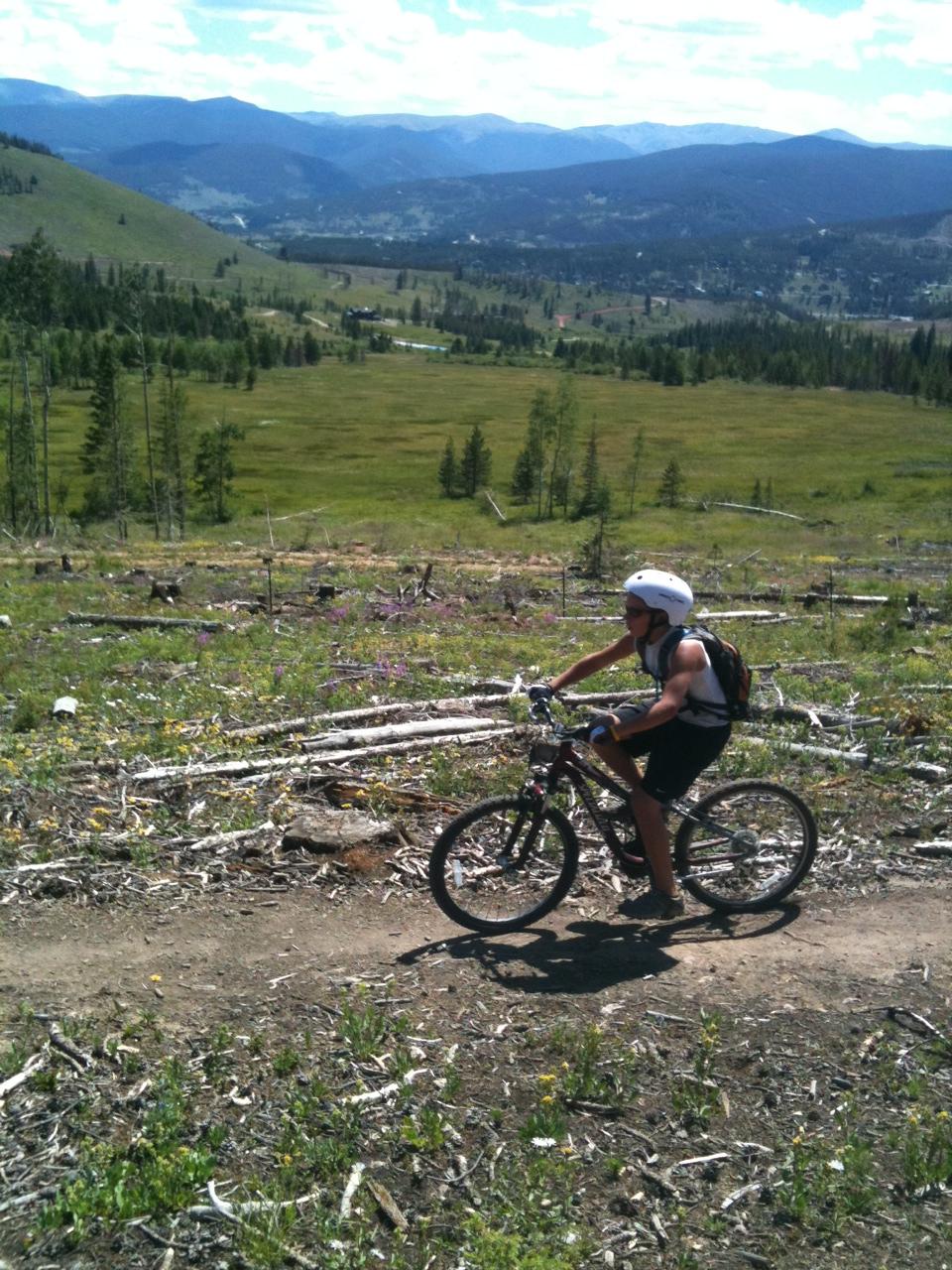 A person riding a mountain bike along a dirt trail in a mountainous landscape. The background features rolling green hills, scattered trees, and a clear blue sky with fluffy white clouds. Wildflowers are visible along the trail, and distant mountains can be seen in the background. Peaks Trail mountain bike trail.