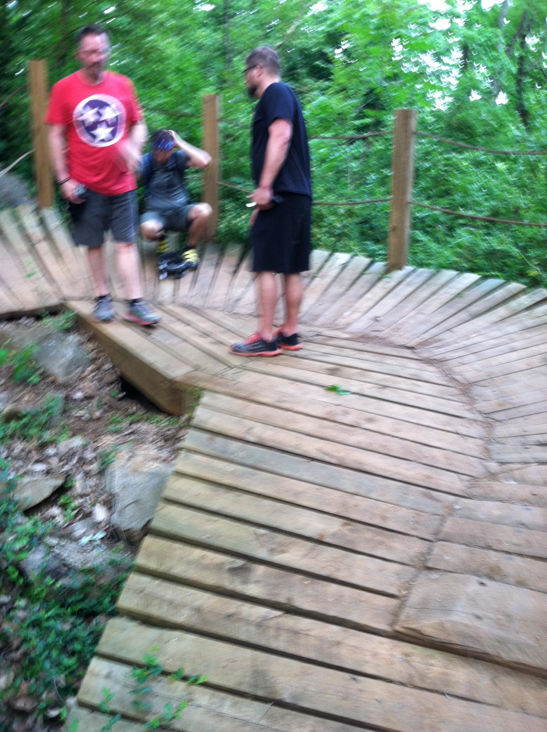 Three people are gathered on a wooden structure surrounded by greenery. One person is sitting down, while the others are standing, engaged in conversation. The ground features a curved wooden pathway, part of a nature trail. The scene appears to be an outdoor recreational area. Ijams Nature Centre mountain bike trail.
