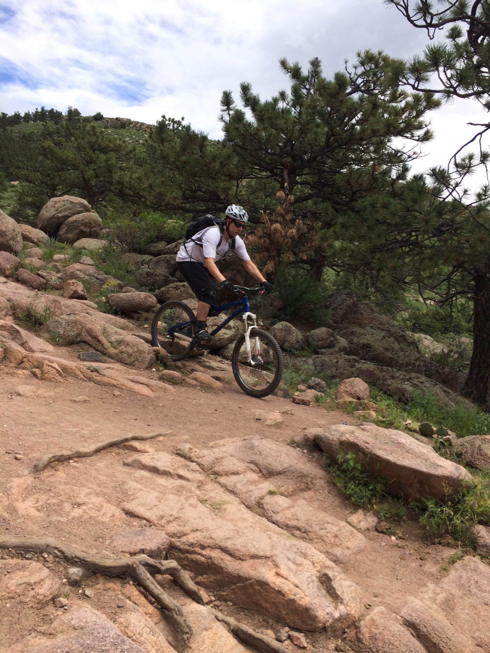 A person riding a mountain bike on a rocky trail surrounded by trees and greenery, with a partly cloudy sky in the background. The terrain features large stones and dirt paths, typical of a mountainous outdoor setting. Hall Ranch mountain bike trail.