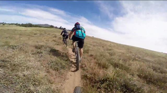 Two mountain bikers riding on a dirt trail through a grassy landscape, with a clear blue sky above and distant hills in the background. Cuyamaca Rancho State Park mountain bike trail.