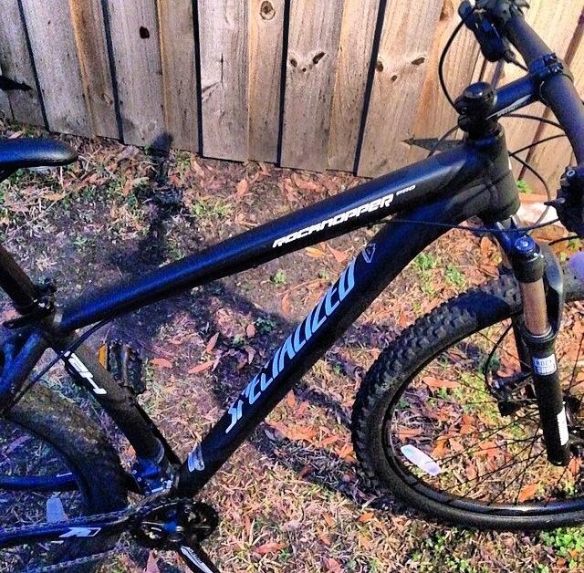 Specialized Rockhopper Pro 29: A close-up image of a black mountain bike with a shiny frame, featuring the brand name "Specialized" and model "Rockhopper" written on the side. The bike is positioned on grass with a wooden fence in the background, showcasing its rugged tires and front suspension.
