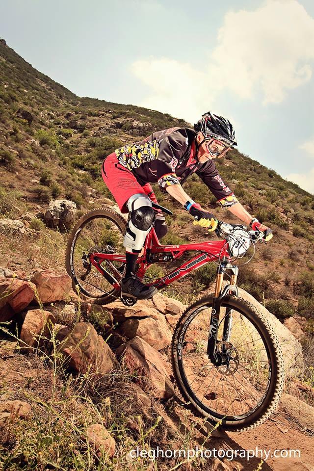 Intense Tracer 275: A mountain biker in protective gear rides a bright red bike over rocky terrain in a hilly landscape. The rider is focused and in motion, showcasing their skills as they navigate the challenging path against a backdrop of greenery and blue sky.