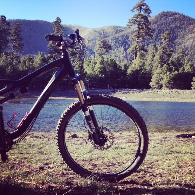 A mountain bike positioned on the shore of a serene lake, surrounded by lush green trees and mountains under a clear blue sky. The bike is angled to showcase its front wheel and handlebars, emphasizing its design against the peaceful natural backdrop. Schultz Creek mountain bike trail.