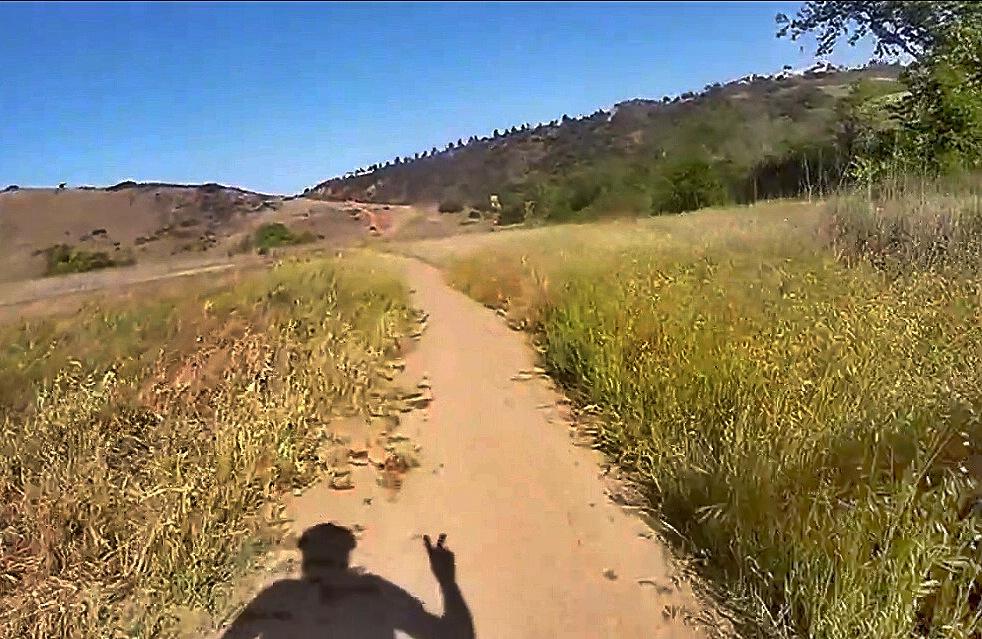 A dirt path winding through a grassy field with hills in the background, under a clear blue sky. A shadow of a person making a peace sign is visible in the foreground. Los Penasquitos Canyon Preserve mountain bike trail.