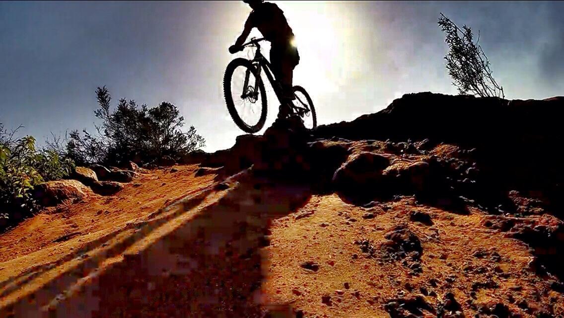 A silhouette of a mountain biker ascending a rocky trail against a bright sun in the background, with shadows cast on the sandy terrain.