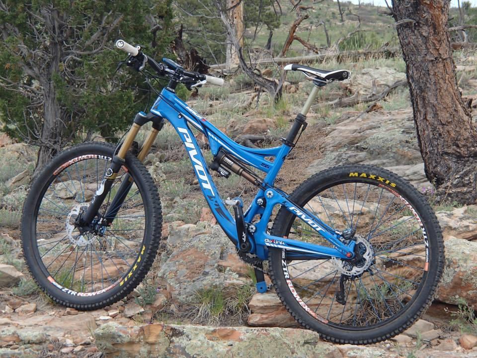 Pivot Mach 6: A blue mountain bike positioned on rocky terrain, surrounded by sparse vegetation and trees. The bike features thick tires suitable for off-road trails and has visible branding on its frame and wheels.