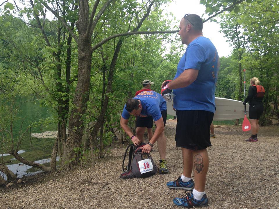 Two men and a woman preparing for an outdoor activity by a body of water. One man is kneeling next to a black bag, possibly organizing gear. The other man stands nearby, wearing a blue shirt, and the woman in the background is dressed in a wetsuit with a paddle nearby. The scene is surrounded by greenery, suggesting a natural, outdoor setting. Ijams Nature Centre mountain bike trail.
