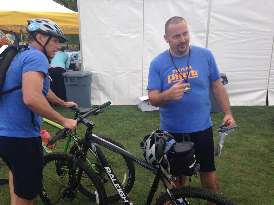 Two men in blue shirts are standing beside their mountain bikes in an outdoor setting. One man is wearing a helmet and is holding the handlebars of a green bike, while the other is gesturing and speaking, possibly giving instructions or information. A white tent and some containers are visible in the background. Ijams Nature Centre mountain bike trail.