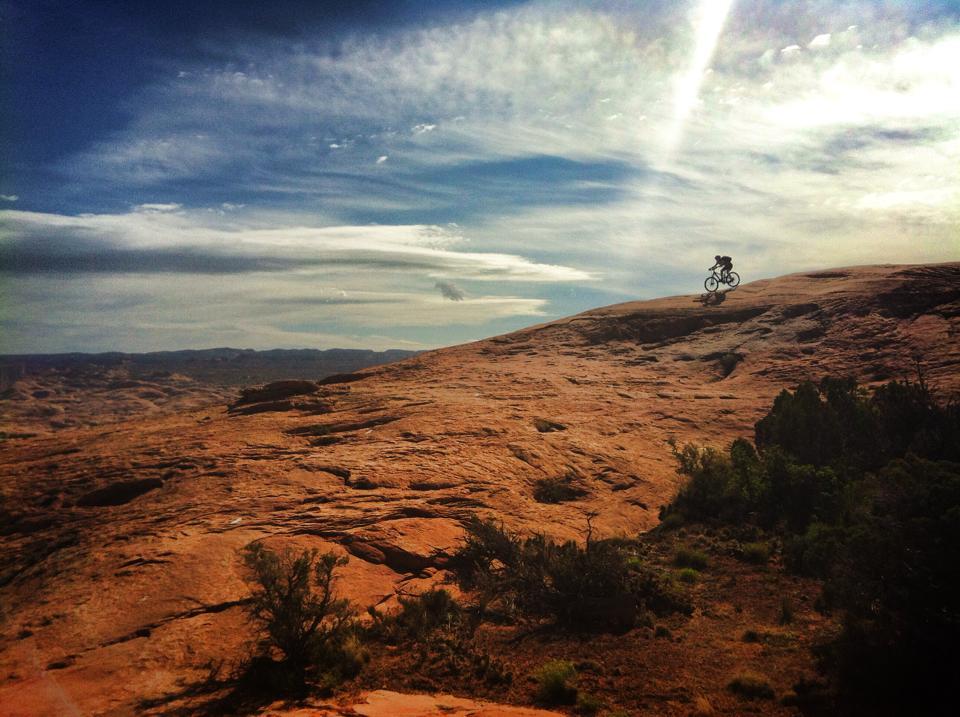 A mountain biker rides along a rocky terrain under a bright, partially cloudy sky, with expansive desert landscape visible in the background. The sun casts a ray of light across the scene, highlighting the textures of the rock and the greenery at the base of the hill. Slickrock mountain bike trail.
