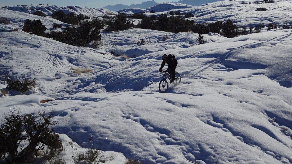 A mountain biker rides through a snowy landscape, navigating over rolling hills covered in white snow, with scattered shrubs and distant mountains visible in the background under a clear sky. Slickrock mountain bike trail.