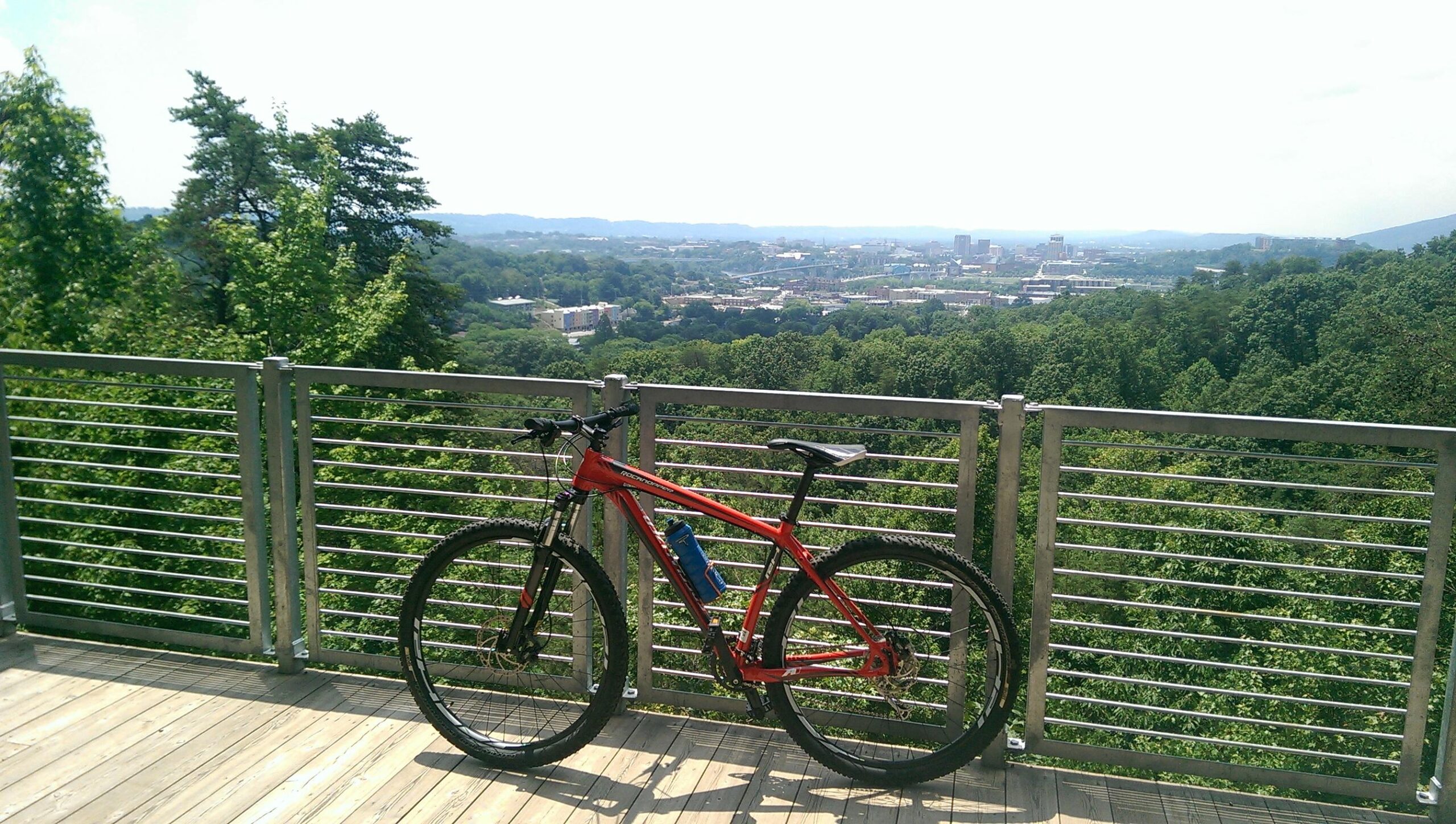Specialized Rockhopper 29: A red mountain bike parked on a wooden platform with a metal railing, overlooking a scenic view of a green landscape and a city in the distance under a bright sky.