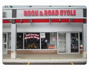 Alt text: Exterior view of a bicycle shop named "Rock & Road Cycle," featuring a bright red sign and a window displaying the text "Full Service Bicycle Shop" along with an "Open" sign. The storefront has a modern design with visible bicycles inside.