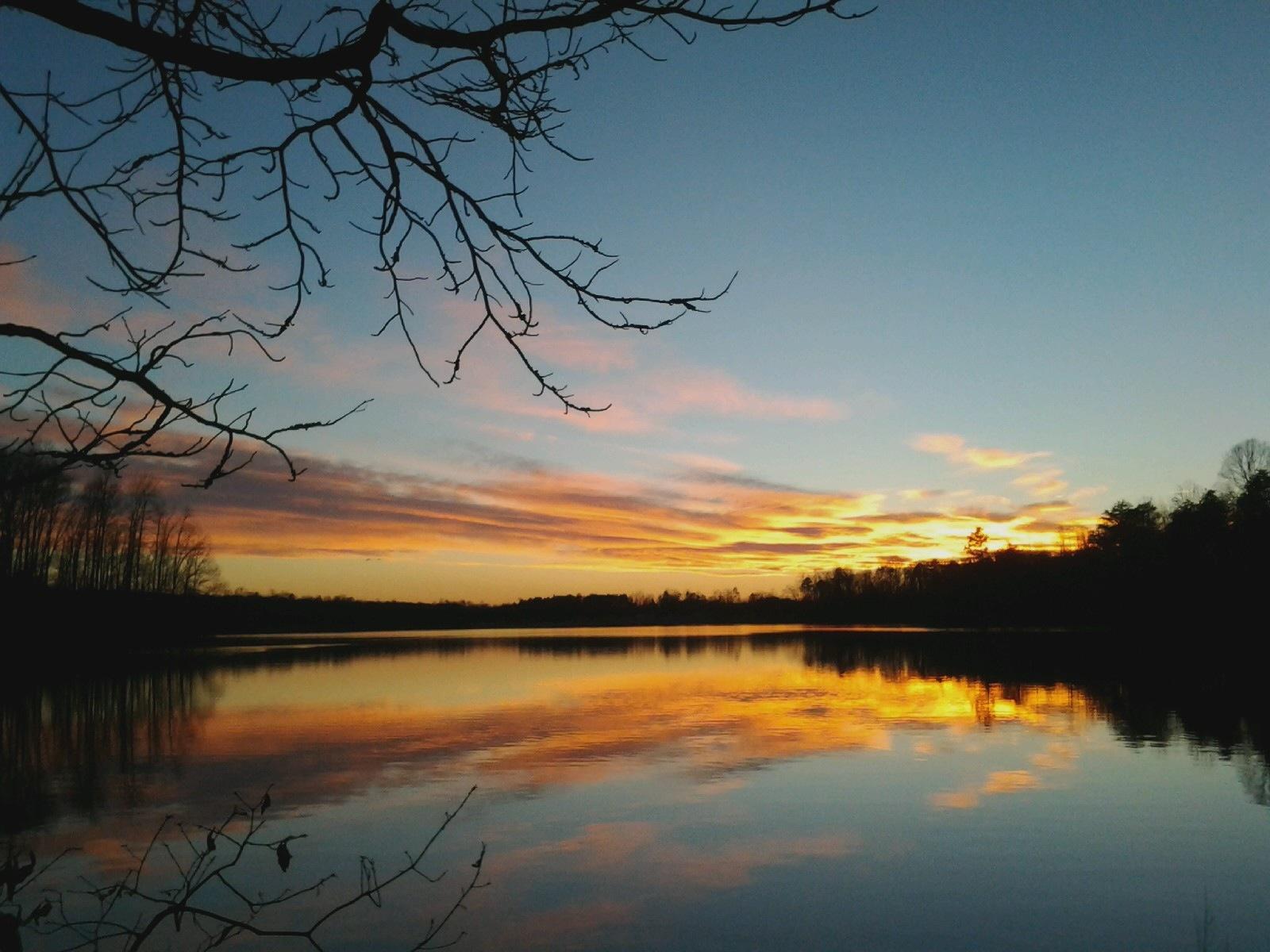 A serene sunset over a calm lake, with vibrant orange and pink hues reflecting on the water. Silhouetted branches frame the top and bottom of the image, while trees line the far shore against a clear blue sky. Salem Lake mountain bike trail.