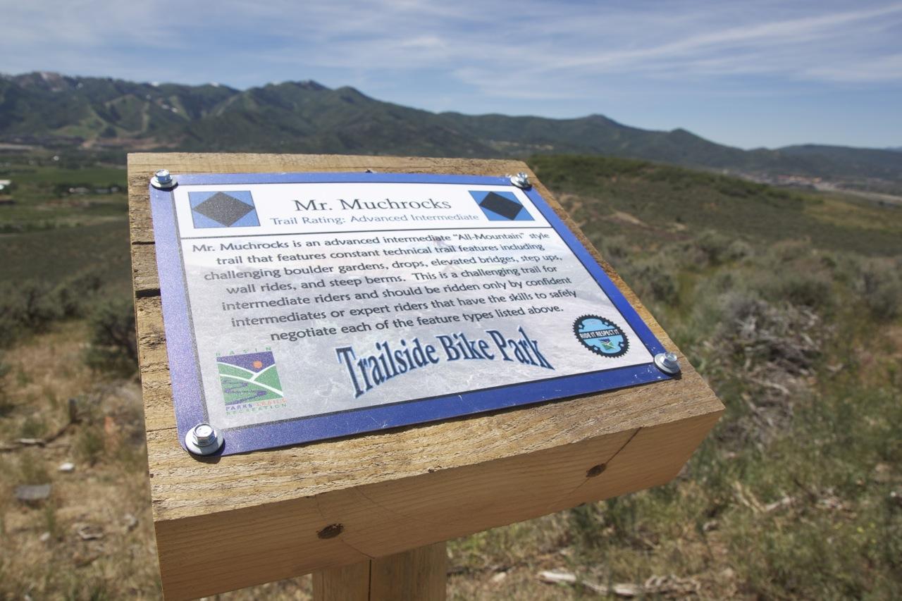 A close-up view of a trail sign for "Mr. Muchrocks" located at the Trailside Bike Park, detailing its advanced intermediate rating. The sign describes the trail's features, including technical elements like boulder gardens, drops, and wall rides, and emphasizes that it should be ridden by confident intermediate or expert mountain bikers. In the background, there are mountains and greenery under a clear blue sky. Trailside Loop And Skills Park mountain bike trail.