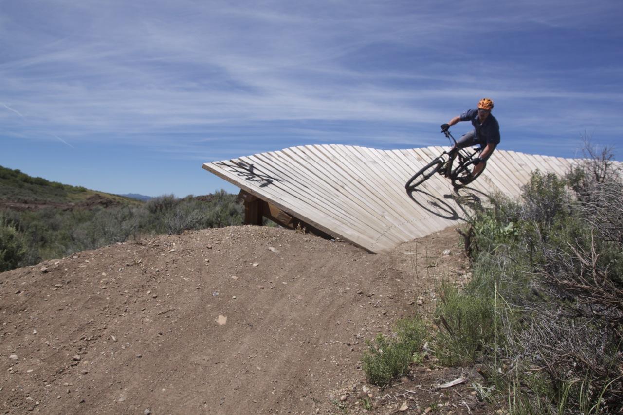 A mountain biker leans into a steep wooden ramp while navigating a dirt trail surrounded by shrubs and underbrush. The sky is clear with light clouds, creating a bright and sunny atmosphere. Trailside Loop And Skills Park mountain bike trail.