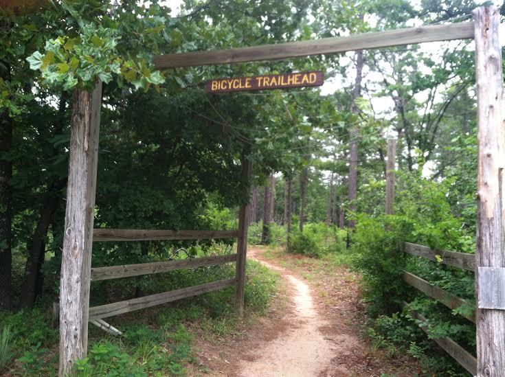 Alt text: A wooden archway displaying the sign "BICYCLE TRAILHEAD" marks the entrance to a dirt path winding through a lush, green forest, flanked by wooden fencing and surrounded by trees. Tyler State Park mountain bike trail.