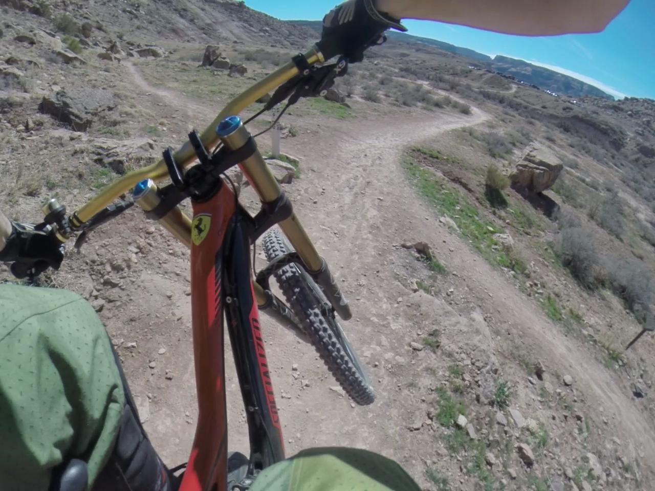 A close-up view of a mountain biker's hands gripping the handlebars of a bike, navigating a winding dirt trail surrounded by rocky terrain and sparse vegetation under a clear blue sky. The bike features a distinctive design with a bright red frame and gold accents. Lunch Loops mountain bike trail.