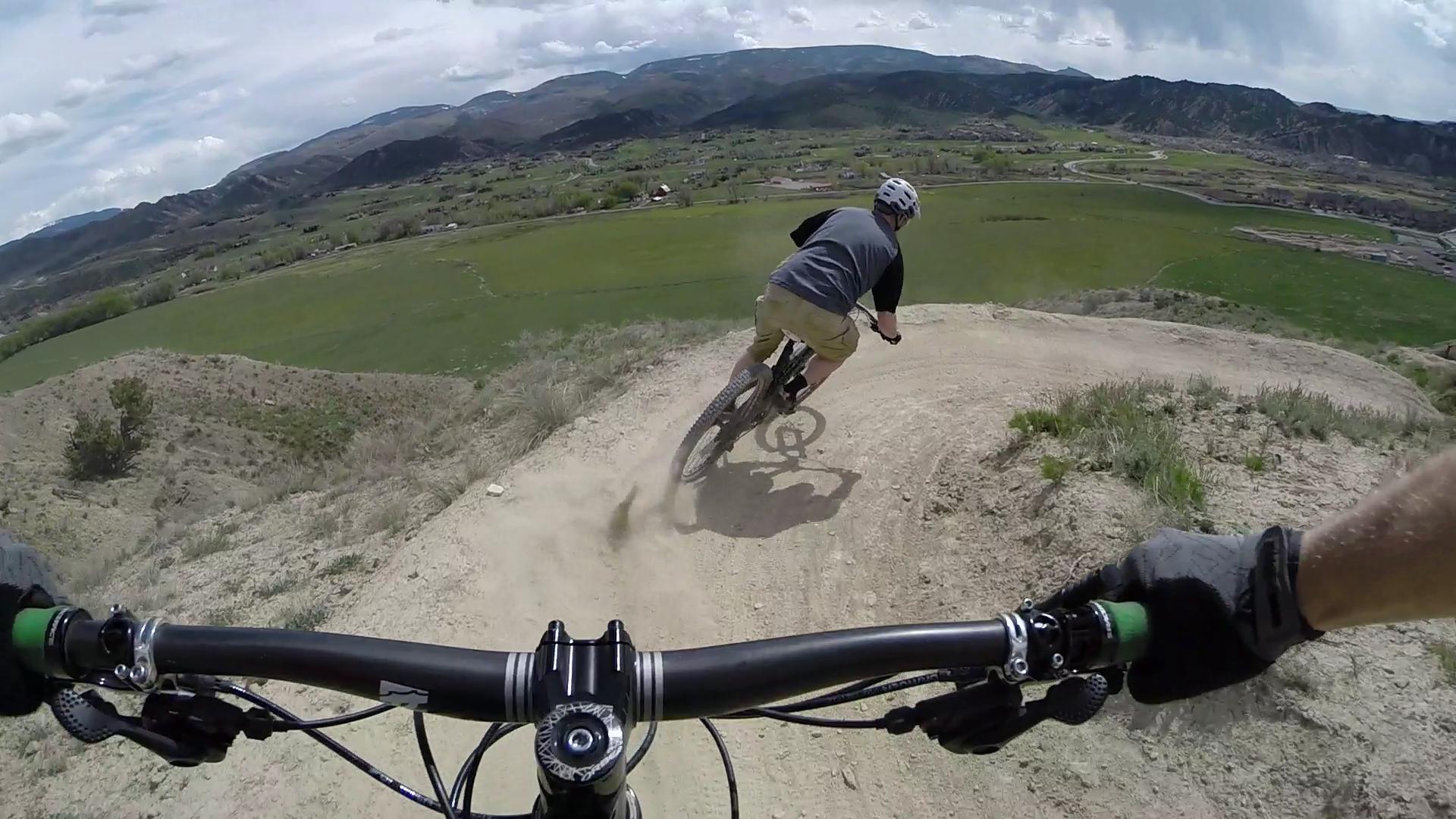 A mountain biker navigating a dirt trail while leaning into a turn, with a panoramic view of green fields and mountains in the background. The perspective includes the biker's handlebars in the foreground, emphasizing the action of the ride. Haymaker Trail mountain bike trail.