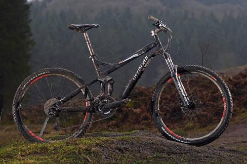 Specialized FSRxc Pro: A mountain bike in a natural outdoor setting, featuring a black frame with red accents, resting on a dirt path surrounded by trees. The bike appears slightly muddy, indicating recent use in rugged terrain.